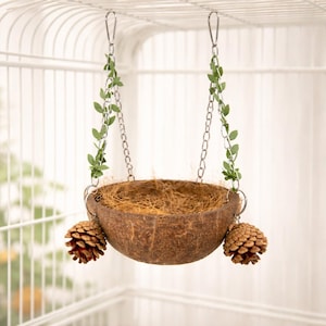 May include: A hanging bird nest made from a halved coconut shell, suspended by metal chains and adorned with faux greenery. Two pinecones are attached to the sides. The nest is designed to be placed inside a birdcage.