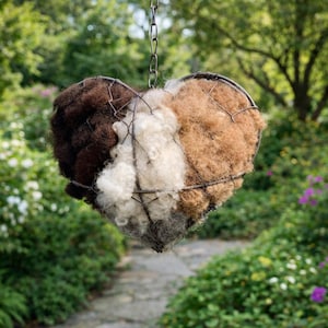 May include: A heart-shaped hanging planter filled with three sections of natural fibres in shades of brown, white, and tan. The planter is suspended by a metal chain, with a wire frame. The background features a blurred garden path and greenery.