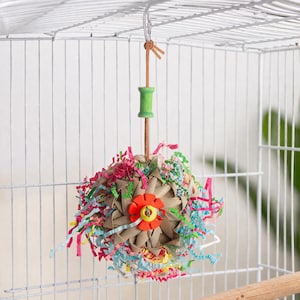 Pu&ograve; includere: Un colorato giocattolo per uccelli appeso all'interno di una gabbia di filo bianco. Il giocattolo &egrave; una palla intrecciata con strisce di carta colorate e un fiore rosso. &Egrave; sospeso da un tassello di legno e una bobina verde.