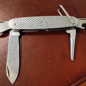 May include: A silver-toned multi-tool pocket knife with a textured handle featuring the letters U.S. and the text CAN OPENER. The tool includes a main blade, a smaller blade, a bottle opener, and a metal bail loop, all constructed from metal.