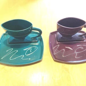 May include: Two ceramic teacups and saucers. One set is teal with white abstract lines, the other is plum with white abstract lines. The cups are square with handles and sit on square saucers.