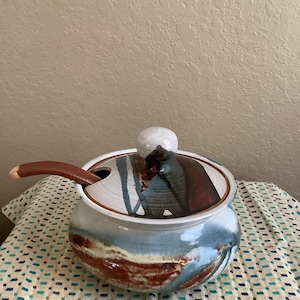 May include: A ceramic soup tureen with a lid and a wooden spoon. The tureen is decorated with abstract patterns in shades of blue, brown, and white.