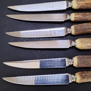 May include: A set of six steak knives with silver-toned metal blades and textured, brown antler-style handles. Each knife features a serrated edge and a metal bolster connecting the blade to the handle. The knives are arranged horizontally.