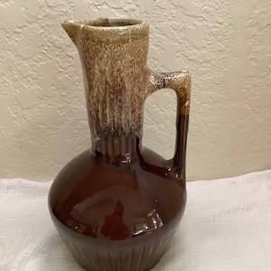 May include: A brown ceramic pitcher with a light brown and white glaze. The pitcher has a wide base and a narrow neck with a spout. The handle is attached to the side of the pitcher.