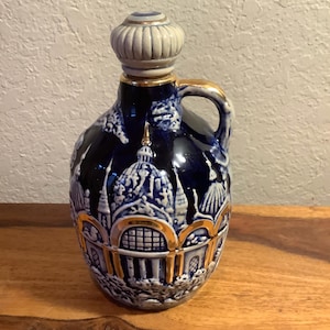 May include: A blue and white ceramic decanter with a gold trim and a detailed design of buildings. The decanter has a handle and a gold-colored stopper.