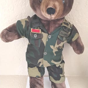 May include: A brown teddy bear wearing a camouflage U.S. Army uniform. The bear has dark brown fur, a lighter brown snout, and button eyes. The uniform includes a jacket with a red and gold patch and the words "U.S. ARMY".