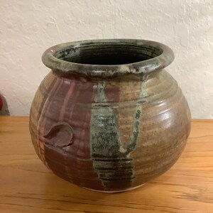Op de afbeelding: Handgemaakte keramische pot met een brede, afgeronde body en een licht uitlopende rand. De pot heeft een mix van aardetinten, waaronder bruin, beige en groen, met een textuur en een klein, decoratief handvat. Een uniek stuk voor de woondecoratie.