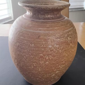 May include: A rustic, brown ceramic vase with a wide, rounded body and a narrow neck. The vase has a textured surface with a speckled appearance, and a slightly flared rim. The color is a warm, earthy tone, suitable for home decor.