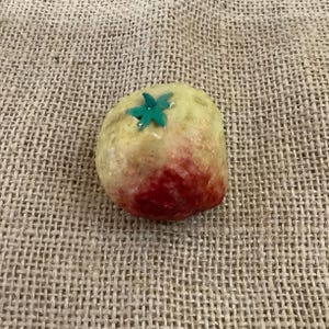 May include: A small, round, decorative object resembling a tomato. The top is light yellow with a green star-shaped accent, and the bottom is a deep red. The object is set against a textured, tan background.