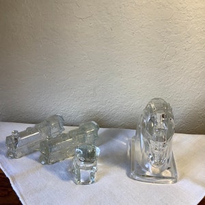 May include: Three clear glass train carriages and a clear glass dog figurine. The train carriages are connected together and the dog is standing on a white surface.