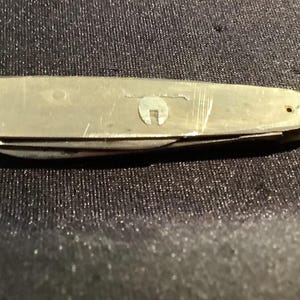 May include: A closed silver-toned pocket knife with a smooth, metallic finish. The handle features a small, circular emblem with a cutout design on the side. The surface of the metal shows light scratches and signs of wear across its length.