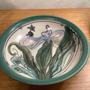 May include: A round ceramic bowl with a teal rim and a white interior. The bowl features a hand-painted illustration of a light purple iris flower with green leaves, a small black starfish, and a black snail on the inner surface of the bowl.