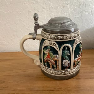 May include: A ceramic beer stein featuring a dark green body with embossed, colorful scenes of figures in traditional attire. It has a cream-colored handle, a decorative base, and a hinged pewter lid with a textured, metallic finish.