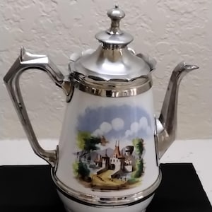 May include: A white porcelain teapot with a silver lid and handle. The teapot has a painted scene of a village with a bridge and a castle. The teapot is sitting on a black surface.