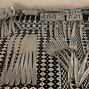 May include: A selection of stainless steel cutlery, including spoons, forks, knives, and a serving spoon, arranged on a black and white patterned tablecloth. The cutlery has a decorative floral design.