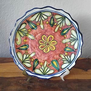Puede incluir: Un plato de cerámica decorativo con borde festoneado. El plato presenta un fondo rojo con un centro floral amarillo y diseños florales y de hojas repetidos en amarillo, verde y blanco. Un borde azul enmarca el diseño.