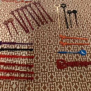 May include: A collection of various cocktail stirrers in red, black, orange, purple, and blue. Some stirrers have circular tops, while others have decorative shapes or text. The stirrers are arranged on a patterned brown and white surface.