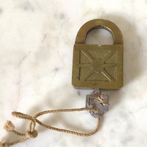May include: A vintage brass padlock with a key attached by a tan rope. The padlock has a square body with a decorative cross pattern and a curved handle. The key is a small, rectangular shape.