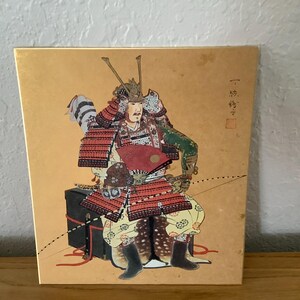 May include: A decorative box featuring a detailed illustration of a samurai warrior in full armor. The artwork showcases a seated figure with a red fan, set against a gold background. Japanese text is visible on the right side of the box.