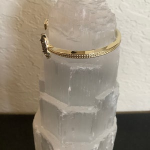 May include: A gold bangle bracelet with a small, dark grey stone set in the centre. The bracelet is on a white selenite crystal.