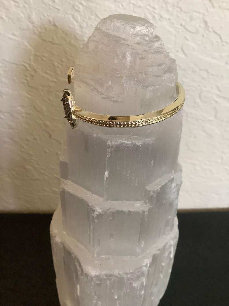 May include: A gold bangle bracelet with a small, dark grey stone set in the centre. The bracelet is on a white selenite crystal.
