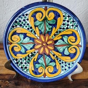 Puede incluir: Un plato de cerámica decorativo con un vibrante diseño floral. El plato presenta un borde azul oscuro y un patrón central de elementos florales amarillos, verdes y naranjas. El diseño es simétrico y detallado, con una mezcla de formas geométricas y orgánicas. El plato se exhibe sobre un soporte de latón.