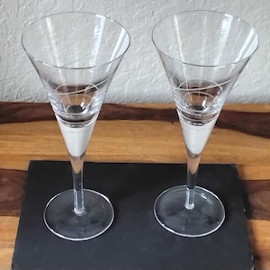 May include: Two clear champagne flutes with a white swirl design. The flutes have a long stem and a wide, conical bowl. The base of the flutes are round. The flutes are on a black surface.