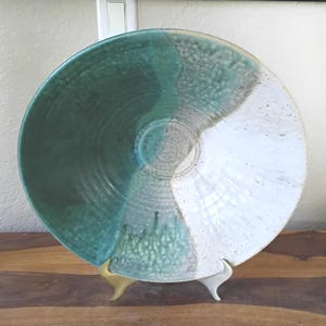 May include: A large, handmade ceramic bowl with a divided design. One half is a deep teal, the other is a speckled white. The bowl is supported by a gold-coloured stand, showcasing its unique, artistic craftsmanship.