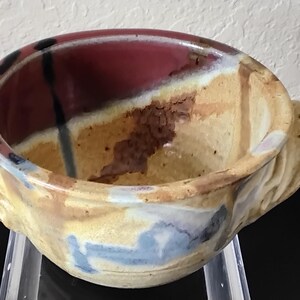 May include: A handmade ceramic bowl with a unique design. The bowl features a combination of colors, including red, yellow, brown, and blue. It has two handles and is suitable for serving food or as a decorative piece.