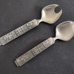 May include: A pair of silver-toned salad servers. One server is a spoon, and the other has a fork-like design. Both have ornate, detailed handles with relief patterns. The servers are displayed on a dark surface.