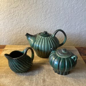 May include: A green ceramic teapot, creamer, and sugar bowl set with a ribbed design. The set is on a burlap surface.