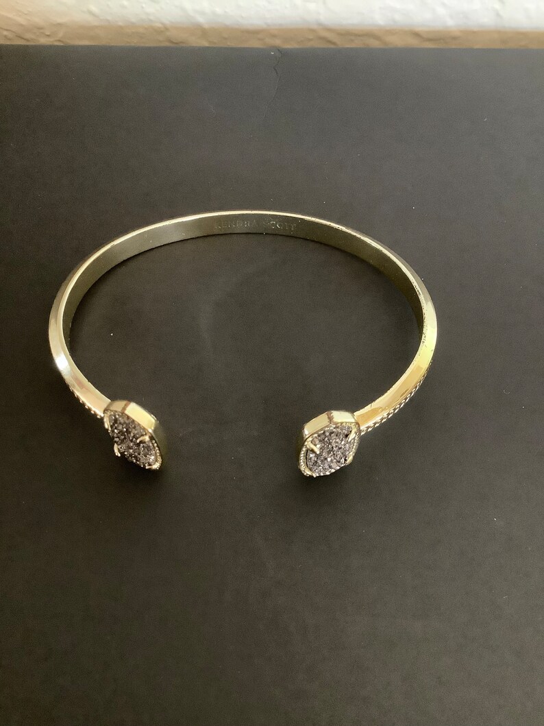 May include: A gold-toned bangle bracelet with a pave-set design on the ends. The bracelet is engraved with the words "Kendra Scott".