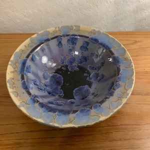 May include: A round ceramic bowl featuring a crystalline glaze with a deep purple and dark blue center. The interior surface is decorated with distinct blue and light blue crystalline patterns that radiate toward the cream-colored rim.
