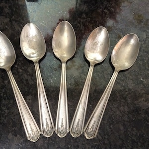 May include: Five silver spoons with ornate designs on the handles. The spoons are arranged in a row, with the handles pointing towards the viewer.