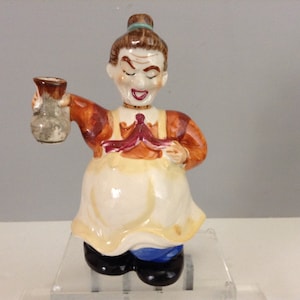 May include: A ceramic figurine of a woman holding a pitcher. She is wearing a brown and yellow dress with a red apron. Her hair is pulled back in a bun and she has a mischievous expression on her face.