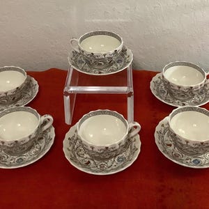 Puede incluir: Un juego de seis tazas y platillos blancos con un diseño floral decorativo gris y azul. Una taza y un platillo se exhiben en un soporte acrílico transparente. Las tazas tienen un borde festoneado.