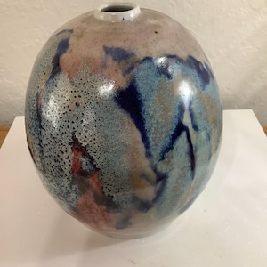 Puede incluir: Un jarrón de cerámica hecho a mano con forma redondeada y una pequeña abertura. El jarrón presenta un esmalte moteado en tonos azules, grises y rosas, con vetas azules más oscuras. La superficie tiene una textura moteada.