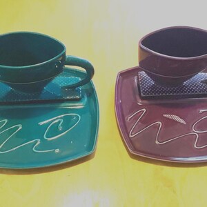 Puede incluir: Dos tazas de té y platillos de cerámica. Una es verde azulado, la otra es de color burdeos intenso. Cada taza se asienta sobre un platillo cuadrado con un diseño abstracto blanco. Las tazas tienen una forma angular única y asas a juego.