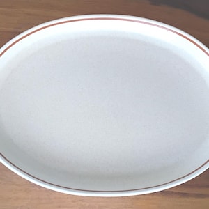 May include: Oval, off-white serving dish with a brown rim and handles. The dish is made of ceramic and is set against a wooden background. This dish is perfect for serving appetizers, side dishes, or desserts.