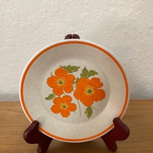 May include: A vintage ceramic plate with a speckled cream background, featuring three orange flowers with green leaves. The plate has an orange border and is displayed on a dark red wooden stand. A classic piece for kitchen decor.