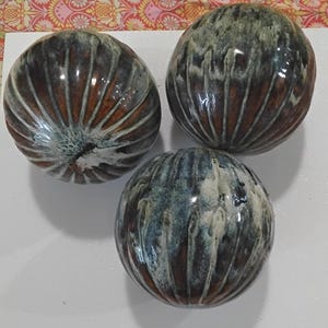 May include: Three spherical ceramic objects with a ribbed, textured surface. The glaze features a blend of deep brown, rust, and mottled grey-blue tones, with white highlights dripping down the vertical ridges of each round decorative piece.