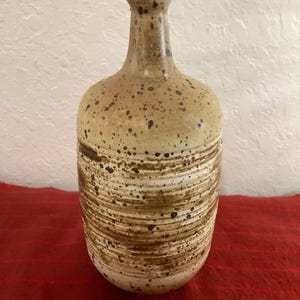 Könnte beinhalten: Eine Keramikvase mit gesprenkeltem, beigefarbenem Körper und schmalem Hals. Die Vase hat horizontale braune Streifen in der Mitte und steht vor einem roten Hintergrund.