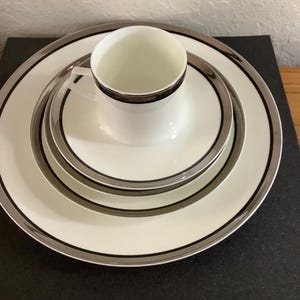 Può includere: Un set di tazza e piattino bianchi con finiture argentate e nere. Il set include un piatto piano, un piatto da insalata e un piatto più piccolo. La tazza è centrata sul piattino e il set è disposto su una superficie scura.