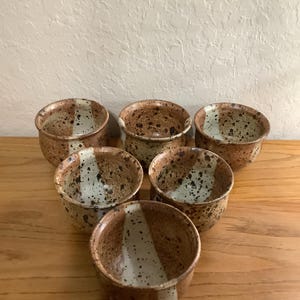 May include: A set of six ceramic bowls featuring a speckled brown and tan glaze with a distinct vertical white stripe running down the center of each piece. The bowls have a rounded shape and a glossy finish, arranged in a triangular formation.