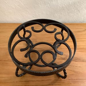 May include: A circular black metal stand featuring a textured outer rim and a decorative interior pattern of interconnected scrolls. The stand is supported by three twisted metal legs that provide elevation and stability for the circular frame.