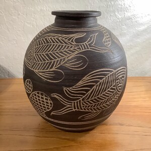 May include: A dark brown, spherical ceramic vase featuring etched, light-coloured line art of two fish swimming in opposite directions, accompanied by circular, grid-patterned motifs. The vase has a short, narrow neck and a flat, circular rim.