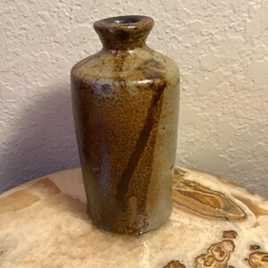 May include: A small, brown ceramic vase with a textured surface and a slightly flared opening. The vase has a light brown glaze with darker brown streaks and a hint of blue at the rim.