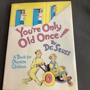 Pode incluir: Um livro amarelo com uma ilustração colorida de um homem empurrando uma cadeira de rodas com outro homem dentro. O título do livro é "You're Only Old Once!" por Dr. Seuss. O texto abaixo do título diz "A Book for Obsolete Children."
