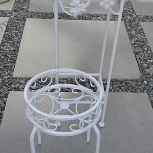 Op de afbeelding: Een witte metalen plantenstandaard met een vierkante bovenkant en een lagere cirkelvormige plantenhouder. De standaard heeft decoratieve bloemaccenten en een traliewerkontwerp op het bovenoppervlak. De standaard is ontworpen om planten te bevatten.