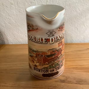 May include: A white ceramic pitcher featuring a colorful graphic of a brewery building and the text Double Diamond Trade Mark Since 1876 Burton on Trent. The cylindrical vessel has a gold-trimmed pouring spout and a smooth, glossy finish.